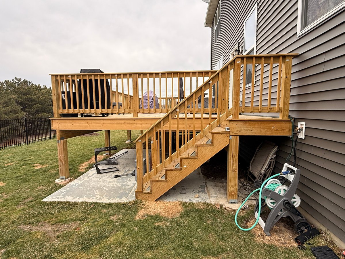 Wooden elevated deck with stairs attached to gray house, featuring railings and concrete patio beneath